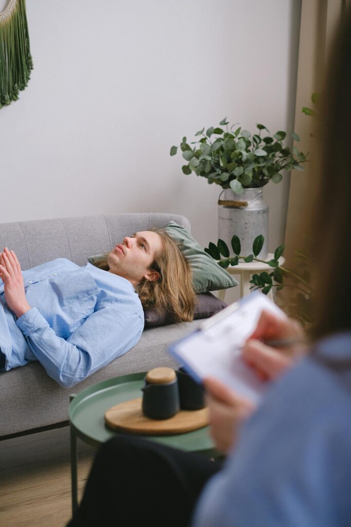 A person in therapy lying on a sofa with a therapist taking notes nearby.
